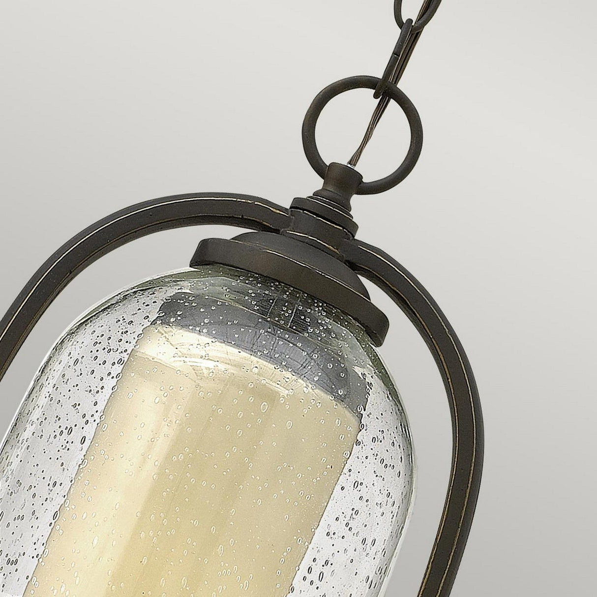 A close-up captures the Quincy Outdoor 1 Light Medium Chain Pendant, showcasing its distinctive seedy outer glass shade with tiny bubbles. Inside, a cream-colored candle-style bulb emits a warm glow. The pendant features an oil-rubbed bronze metal frame, highlighted by its elegant arched design and convenient hanging hook.