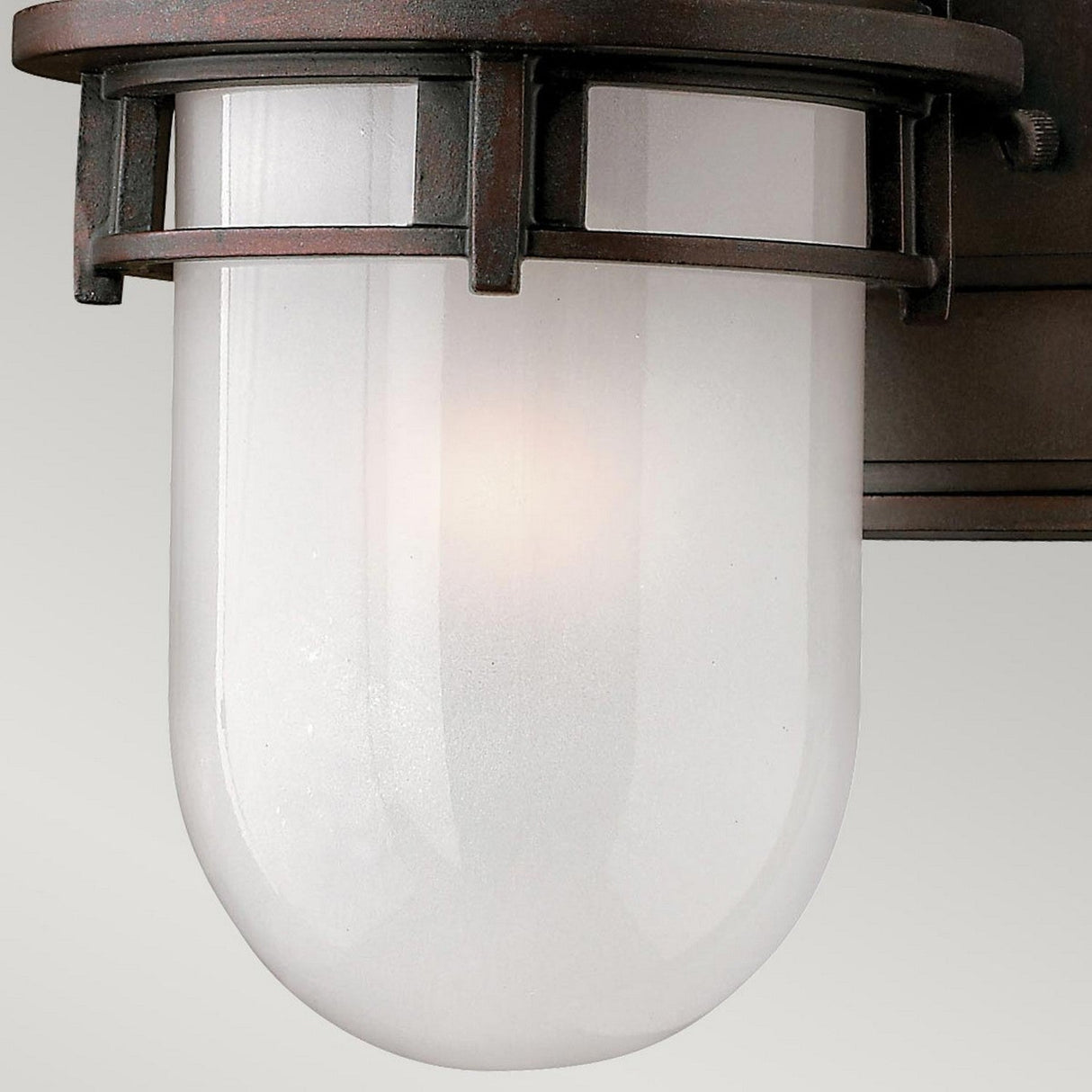 A close-up of the Reef Mini Outdoor 1 Light Lantern - Bronze showcases its industrial-style, wall-mounted design. It features a cylindrical frosted glass shade encased in a dark bronze metal frame made of cast aluminum. With an IP44 rating, this outdoor lantern emits a gentle glow through the glass, providing subtle illumination.