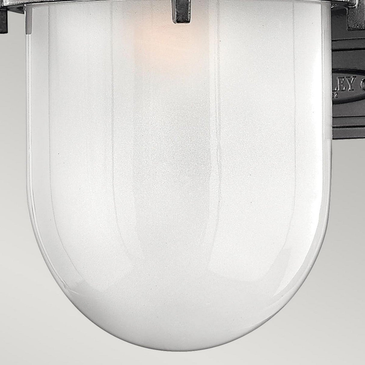 A close-up of the Reef Outdoor Small 1 Light Lantern - Grey features a frosted glass light fixture with a cast aluminum frame. The bulb casts a soft glow through its translucent surface, creating a warm ambiance. This fixture is mounted against a plain background.