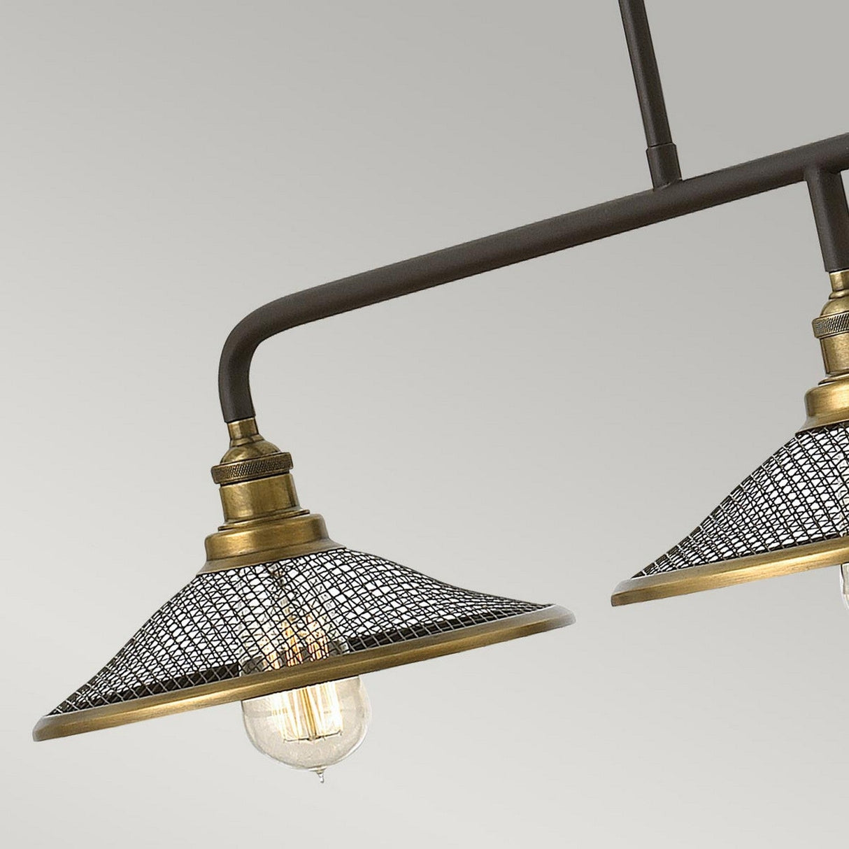 A close-up of the Rigby 3 Light Island Chandelier - Bronze & Brass showcases its mesh-covered lampshades with an antique brass finish. The bulbs emit a glimmering light through the mesh, blending industrial and vintage styles on a dark metal frame, against a subtle gray backdrop.