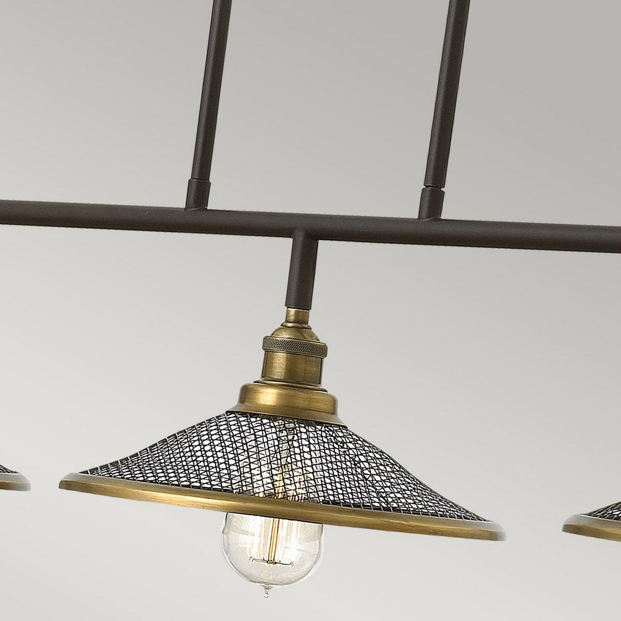 A detailed view of the Rigby 3 Light Island Chandelier - Bronze & Brass showcases its black mesh shade. The visible bulb beneath the mesh casts a warm glow, highlighting the fixture's industrial and vintage design with clean lines against a simple backdrop.