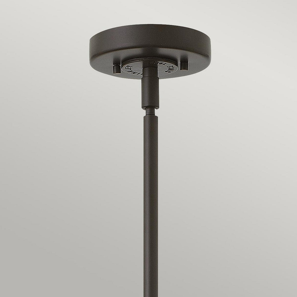 A close-up view of the Rigby 1 Light Pendant in bronze and brass, featuring a sleek cylindrical rod descending from a circular base. The industrial design of this fixture pops against a gradient background of light gray.