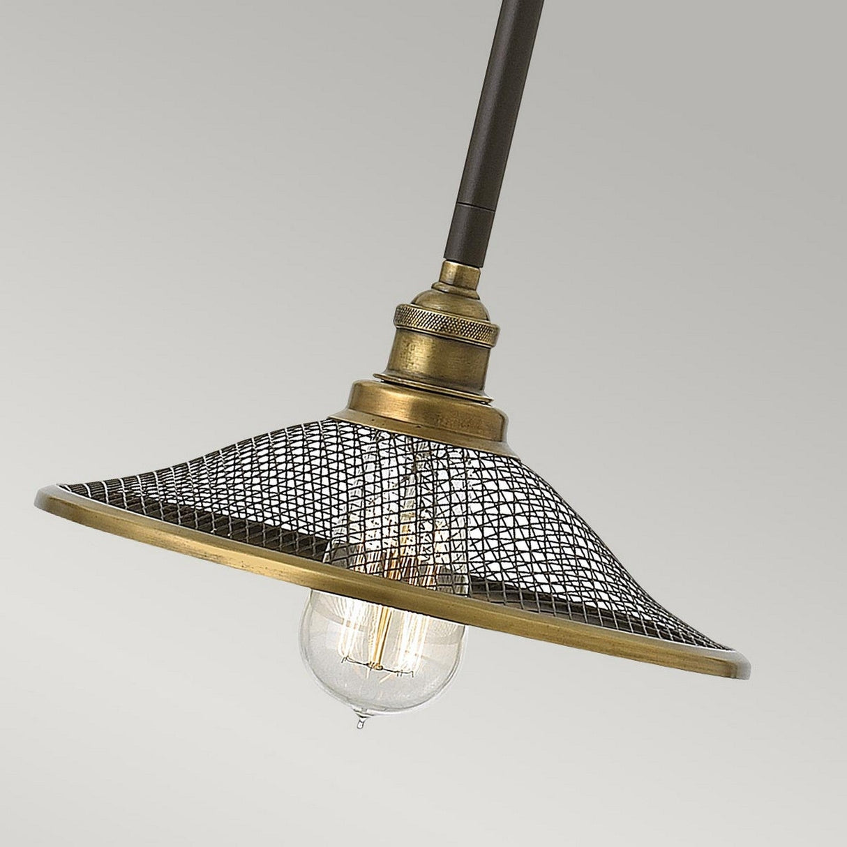 The Rigby 1 Light Pendant in Bronze & Brass features a vintage-style design with a metal mesh shade and an exposed clear bulb, highlighted against a plain gray background. Its brass finish enhances the fixture's industrial lighting appeal, offering a striking blend of vintage charm and modern functionality.