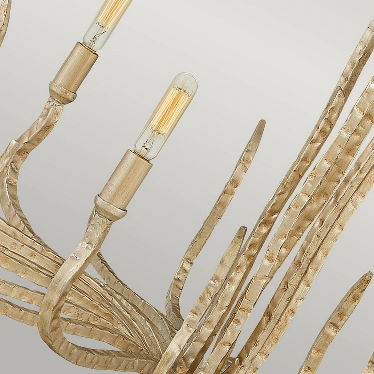 A detailed view of the Spyre 6 Light Chandelier in champagne gold reveals its curved, textured metal rods that mimic branches. The fixture's elegant and organic design is highlighted by two visible cylindrical light bulbs.
