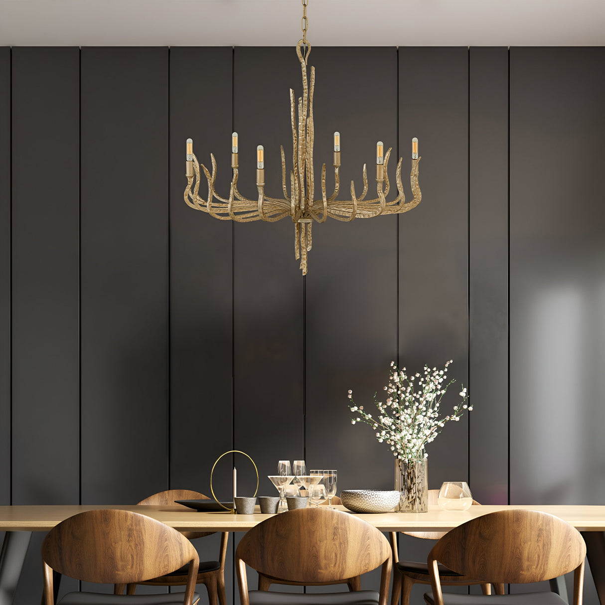 A modern dining room showcases a long wooden table accompanied by four chairs, illuminated by the stunning Spyre 6 Light Chandelier in Champagne Gold. The table is decorated with a vase of white flowers and elegant glassware, set against a dark paneled wall for an added touch of sophistication.