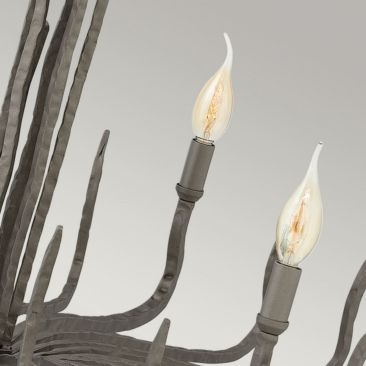 Close-up of the Spyre 6 Light Chandelier in Metallic Matte Bronze with two lit candle-shaped bulbs, showcasing its industrial edge and rustic charm. The chandelier features curved arms against a neutral background.