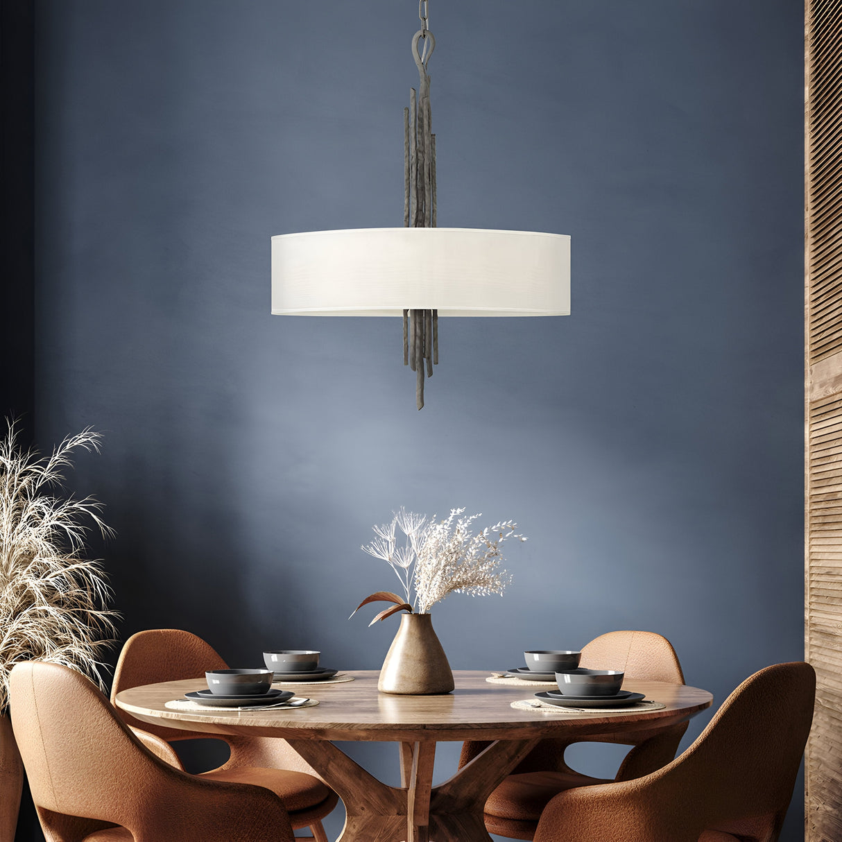 A cozy dining area showcases a round wooden table paired with four brown chairs. Suspended above the table is the Spyre 6 Light Pendant in Metallic Matte Bronze, amplifying the industrial charm with its sculptural design. The centerpiece of the table is a ceramic vase filled with dried flowers, set against a backdrop of a blue wall and wooden paneling.