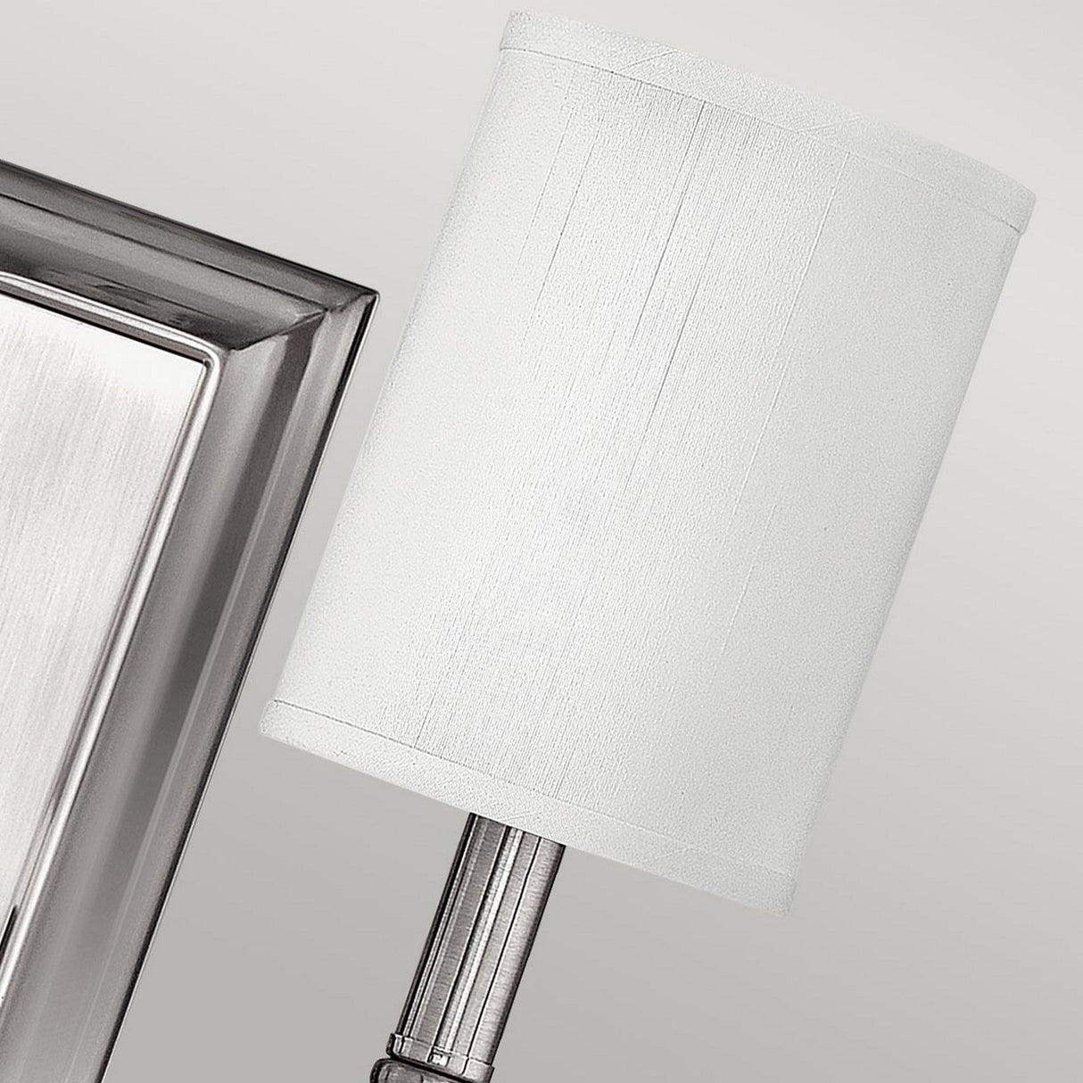 Close-up of the Sussex 2 Light Wall Light - Brushed Nickel, featuring a cylindrical white lampshade. The fixture exudes contemporary elegance with its brushed nickel finish and is mounted on a rectangular backplate against a light gray background.