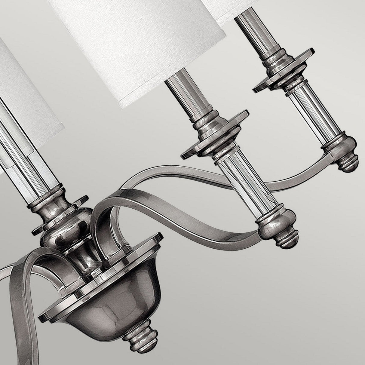 Close-up of the Sussex 5 Light Chandelier - Brushed Nickel, featuring metallic arms and cylindrical lampshades. Its brushed nickel finish exudes traditional charm with sleek curves and intricate detailing, offering a contemporary look against a neutral background.