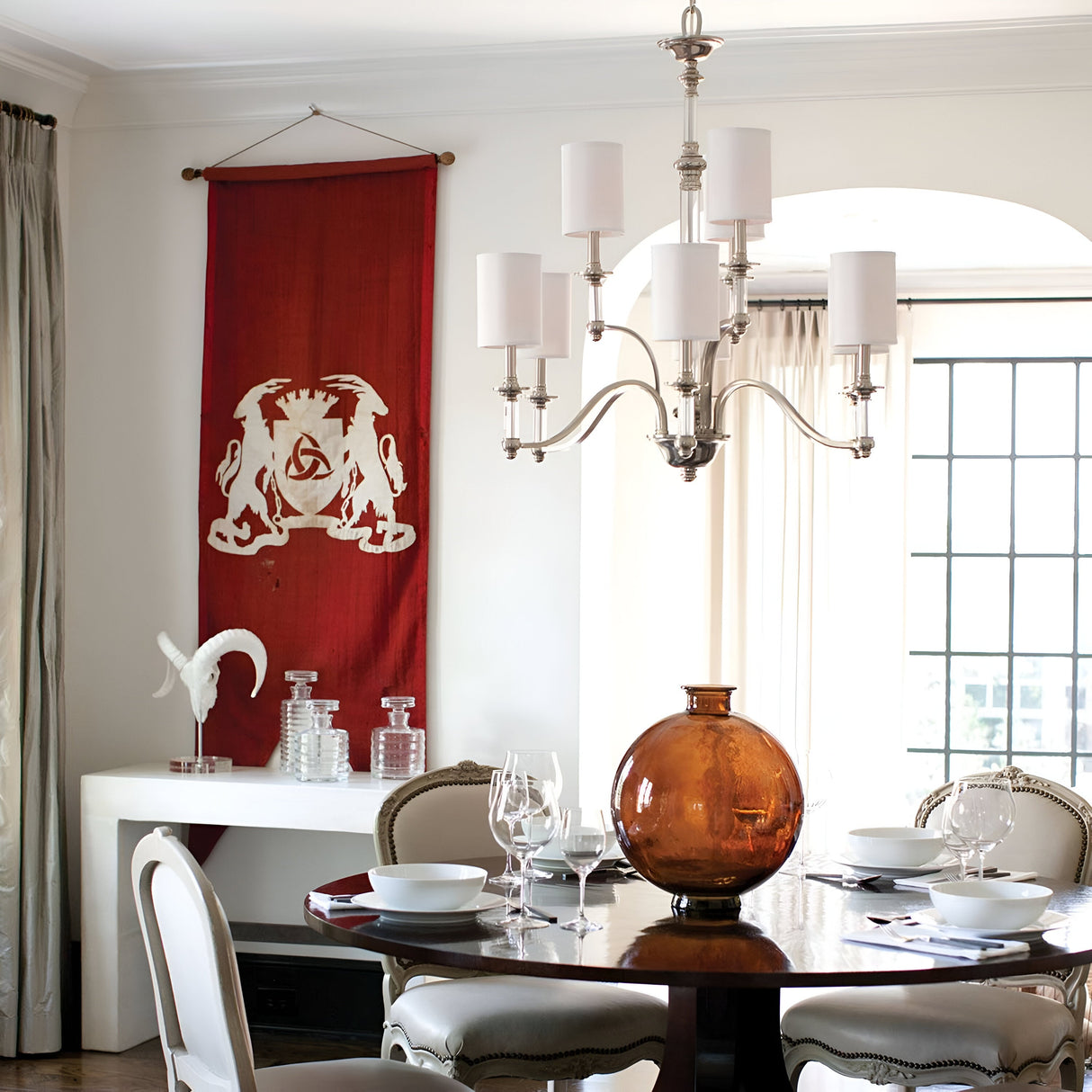 In the dining room, a round table is elegantly set with white plates and wine glasses, while a large amber-colored glass vase takes center stage. A Sussex 9 Light Chandelier in brushed nickel provides graceful illumination overhead, and a red wall hanging with an intricate design decorates the area near the window.