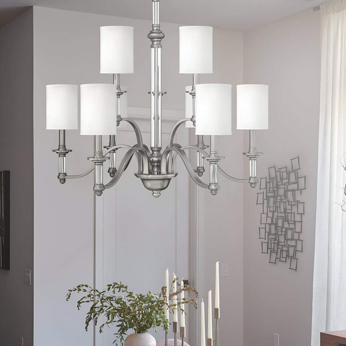 A modern dining room showcases a Sussex 9 Light Chandelier in brushed nickel, elegantly suspended above a sleek dining table. The fixture harmonizes beautifully with the decorative wall piece, while a centerpiece of green foliage and several lit candles illuminate the space under bright natural light.