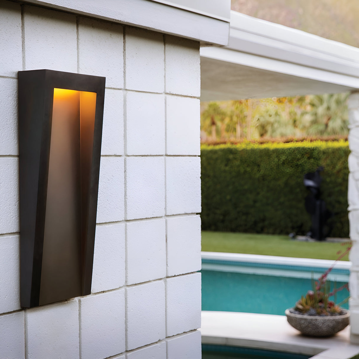 A Taper Outdoor LED Large Wall Lantern in black casts light on a white brick wall. In the background, a swimming pool is bordered by lush greenery, including a manicured hedge and a potted plant. The ambiance is serene and contemporary, highlighting sophisticated outdoor lighting.