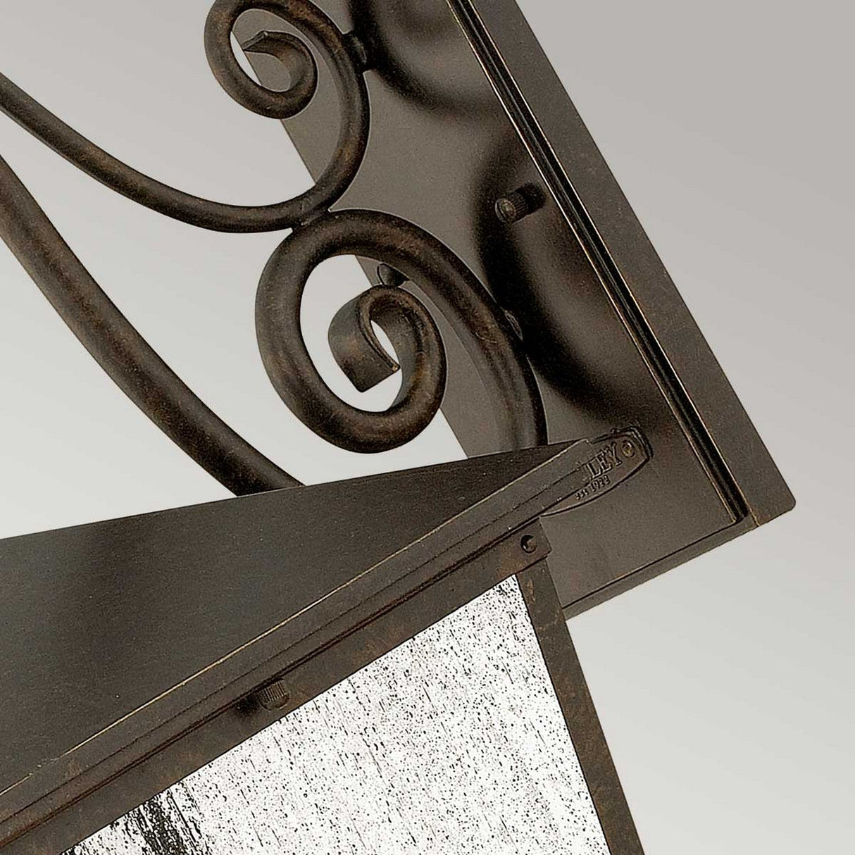 Close-up of the Trellis Outdoor 4 Light Large Wall Lantern in bronze, featuring a black metal wall bracket with ornate spiral detailing and supporting a seedy glass panel. The image highlights the lantern's intricate design against a plain gray background.