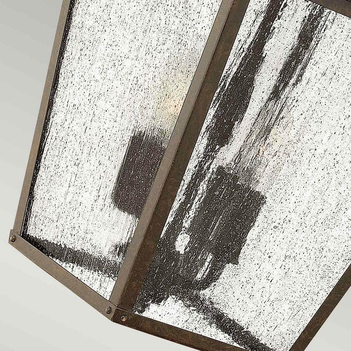 A close-up view of the Trellis Outdoor 4 Light Large Wall Lantern - Bronze reveals its seedy glass panels, each with a speckled pattern that partially shows the lightbulbs inside. The angle highlights its geometrical design, making it perfect for outdoor lighting.