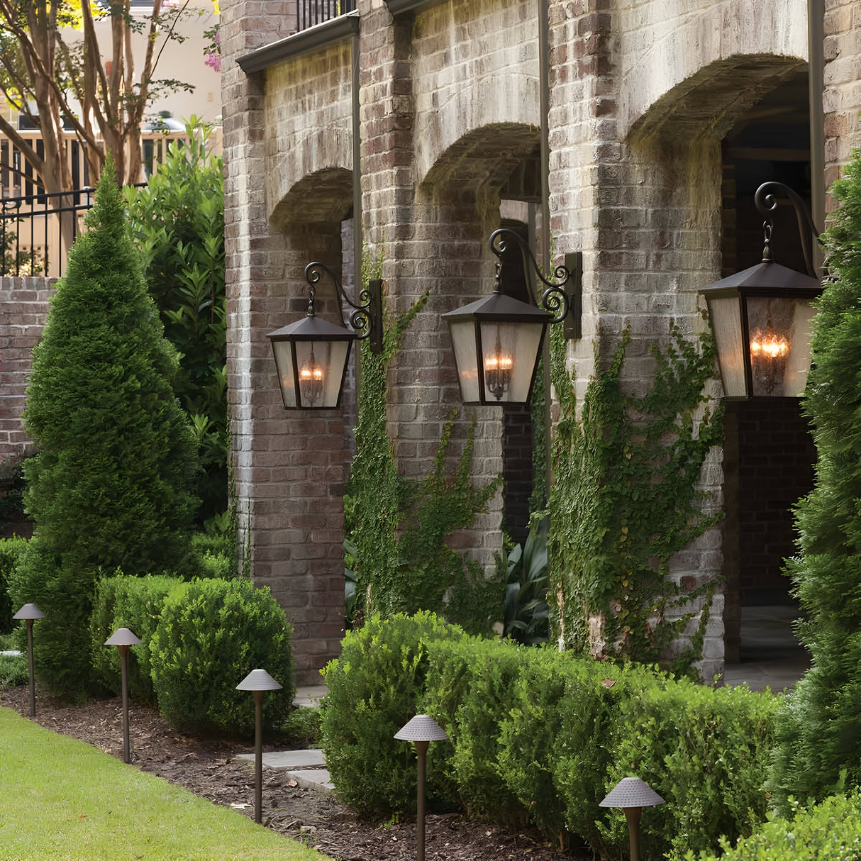 In front of a brick building, a picturesque garden with neatly trimmed bushes and conical trees is enhanced by the Trellis Outdoor 4 Light Large Wall Lanterns in bronze, hanging from arches and casting a warm glow. The vintage-style lights, featuring seed glass panels, add character to the scene, while pathway lighting guides along the grassy area.