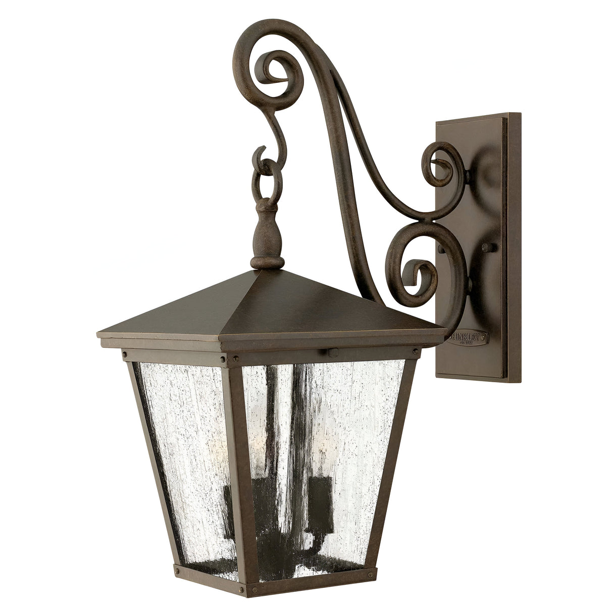 The Trellis Outdoor 3 Light Medium Wall Lantern in bronze showcases a vintage-style design with a metal frame featuring elegant scrollwork. It includes a tapered square glass enclosure crafted from seedy glass, all mounted on a decorative bracket.