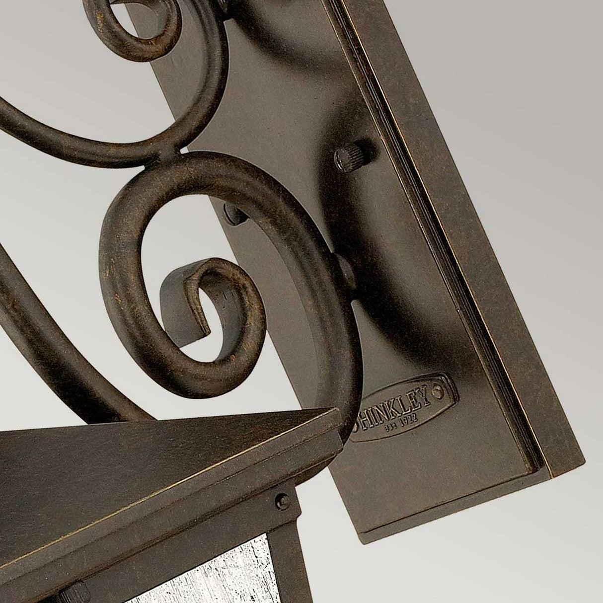 Close-up of the Trellis Outdoor 3 Light Medium Wall Lantern - Bronze, featuring brown metal with ornate scrollwork and seedy glass. A small plaque reads Hinkley Est. 1922. The bronze finish stands out against a simple gradient from light gray to white in the background.