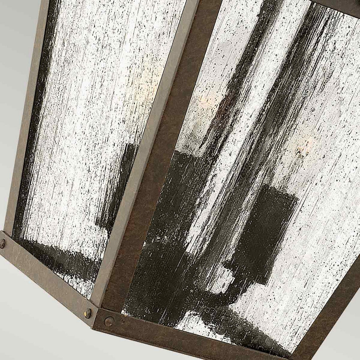 A close-up of the Trellis Outdoor 3 Light Medium Wall Lantern in bronze reveals its rustic charm. The seeded glass panels feature a textured, streaked look, and the fixture is artistically hung at an angle to emphasize its metal frame and bulb holders.