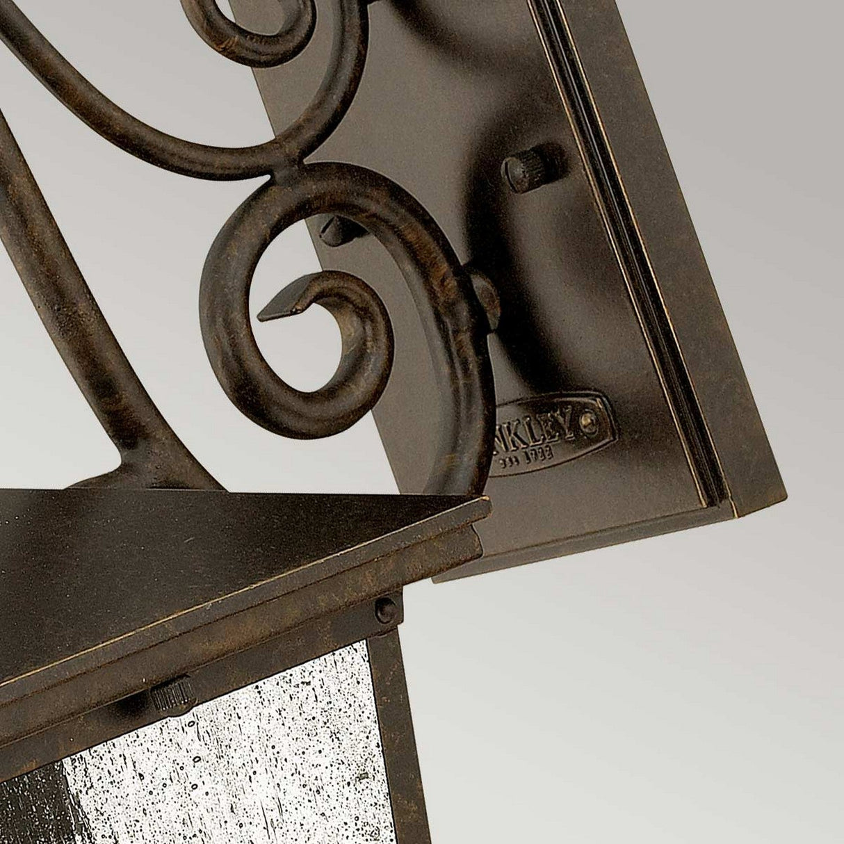 Close-up of the Trellis Outdoor 1 Light Small Wall Lantern in bronze, showcasing its vintage-style intricacies with scrollwork and a weathered finish. The design includes seedy glass panels set among geometric and curved elements, creating a classic, ornate look. Ideal for outdoor lighting, it stands out beautifully against a soft gradient gray background.
