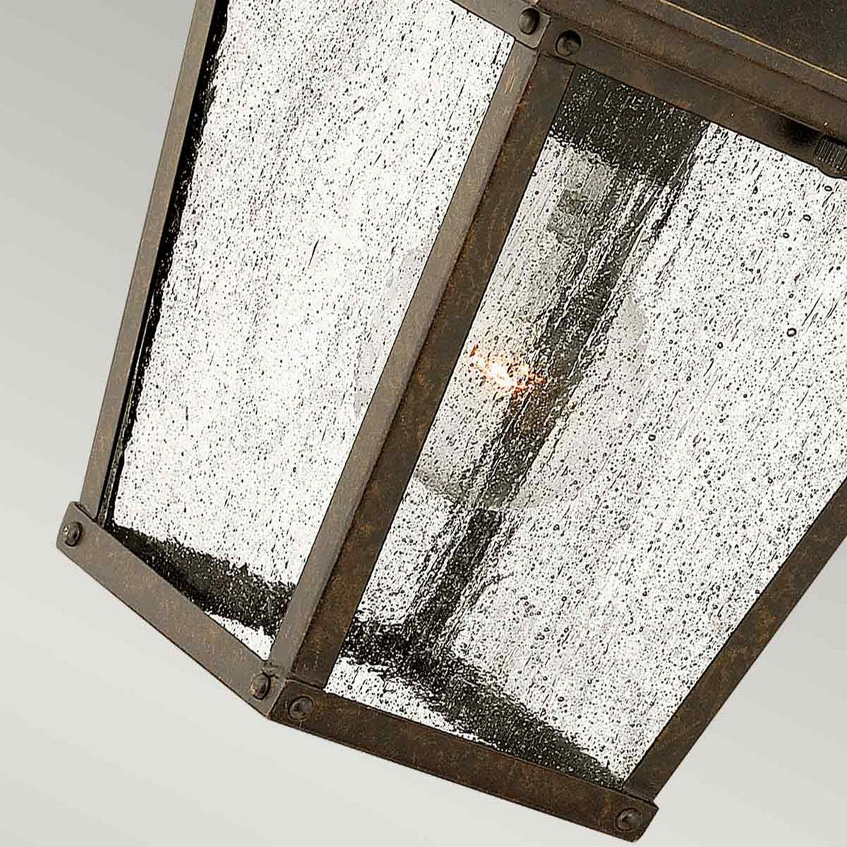 A close-up of the Trellis Outdoor 1 Light Small Wall Lantern in bronze reveals its rustic metal construction and seedy glass panels. Suspended against a light gray background, it elegantly highlights the light bulb within, enhancing the charm of any outdoor lighting arrangement.