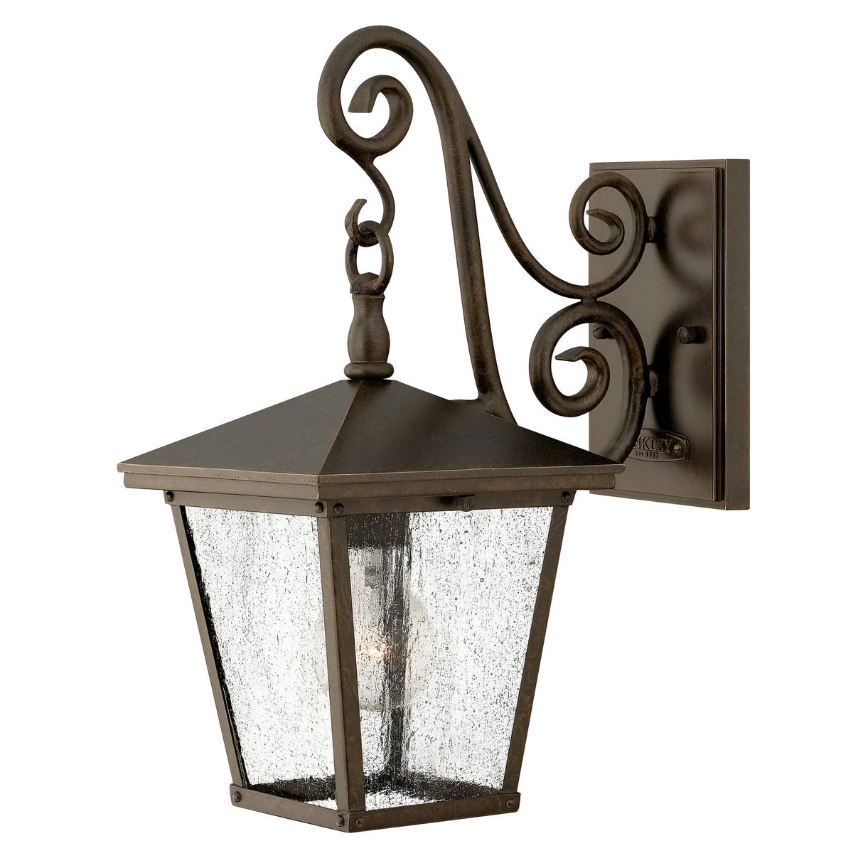 The Trellis Outdoor 1 Light Small Wall Lantern in bronze offers a vintage design with seedy glass panels and decorative scrollwork. It includes a single light bulb inside, mounted on a rectangular base.
