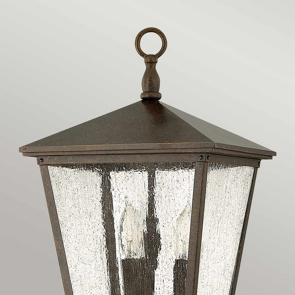 A close-up of the Trellis Outdoor 3 Light Large Pedestal - Bronze, a vintage-style outdoor lighting fixture showcasing a dark metal frame and textured glass pane. This lantern, ideal for illuminating garden paths, boasts a pyramid-shaped top and a decorative loop finial. Inside, three candle-like bulbs emit a warm glow against the plain light gray background.
