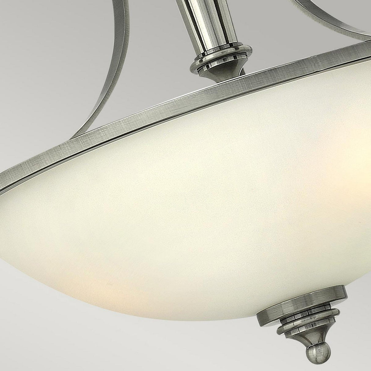 A close-up of the Truman 3 Light Semi Flush in Antique Nickel showcases its frosted glass dome shade. The elegant metal frame gracefully curves above the dome, enhanced by an etched opal glass finish, and a decorative finial hanging below. The light is off, set against a plain gray background.