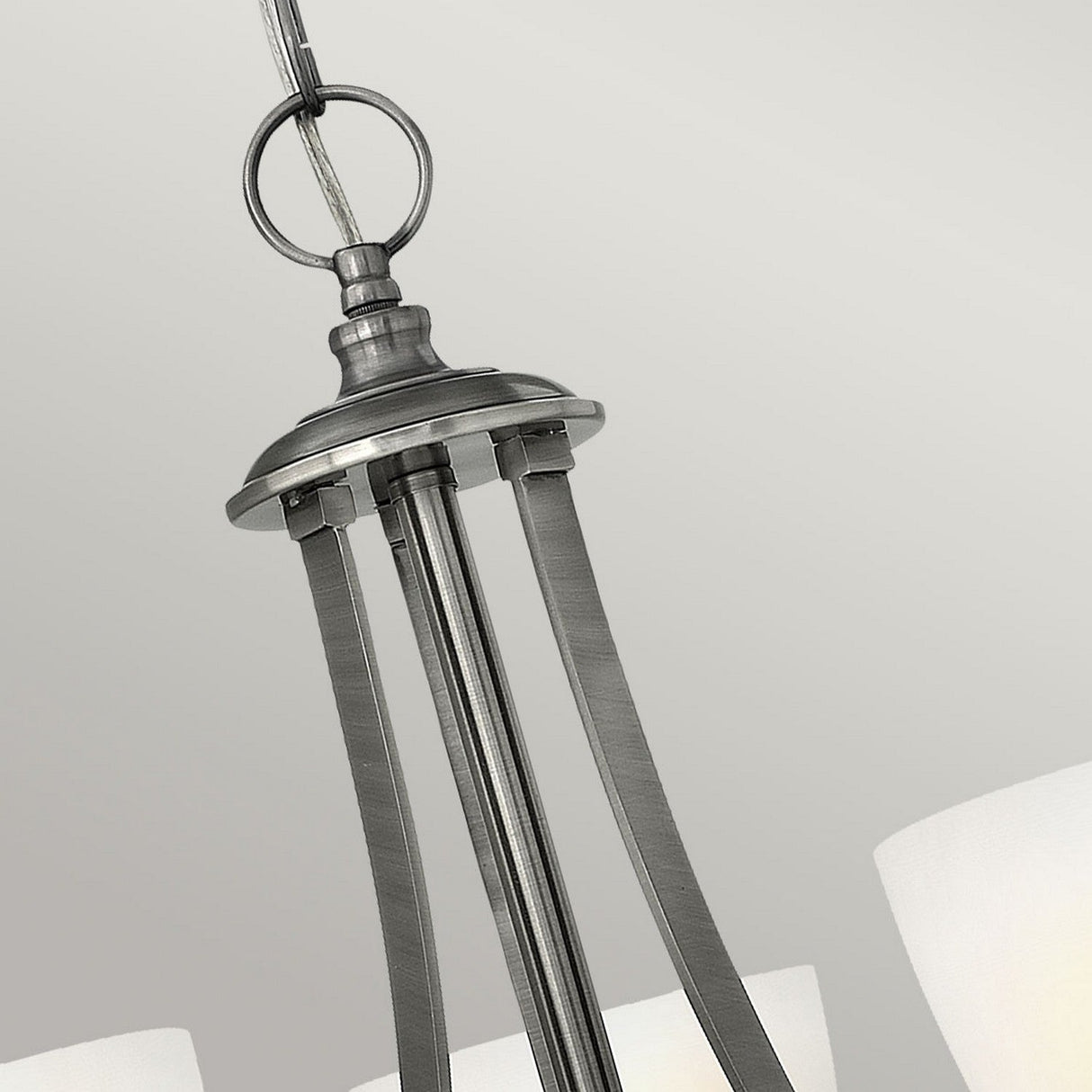 A close-up view of the Truman 3 Light Chandelier - Antique Nickel showcases its modern design, complete with gracefully curved metal arms and etched opal glass shades. The fixture is elegantly suspended from a chain against a neutral gray backdrop.