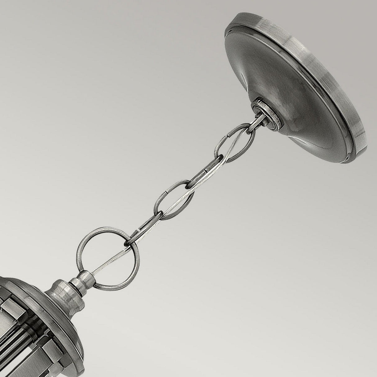 A close-up of a metallic chain connecting to the base of the Truman 5 Light Chandelier in an Antique Nickel finish. The fixture showcases an industrial aesthetic with its brushed metal surface, set against a neutral light gray background.