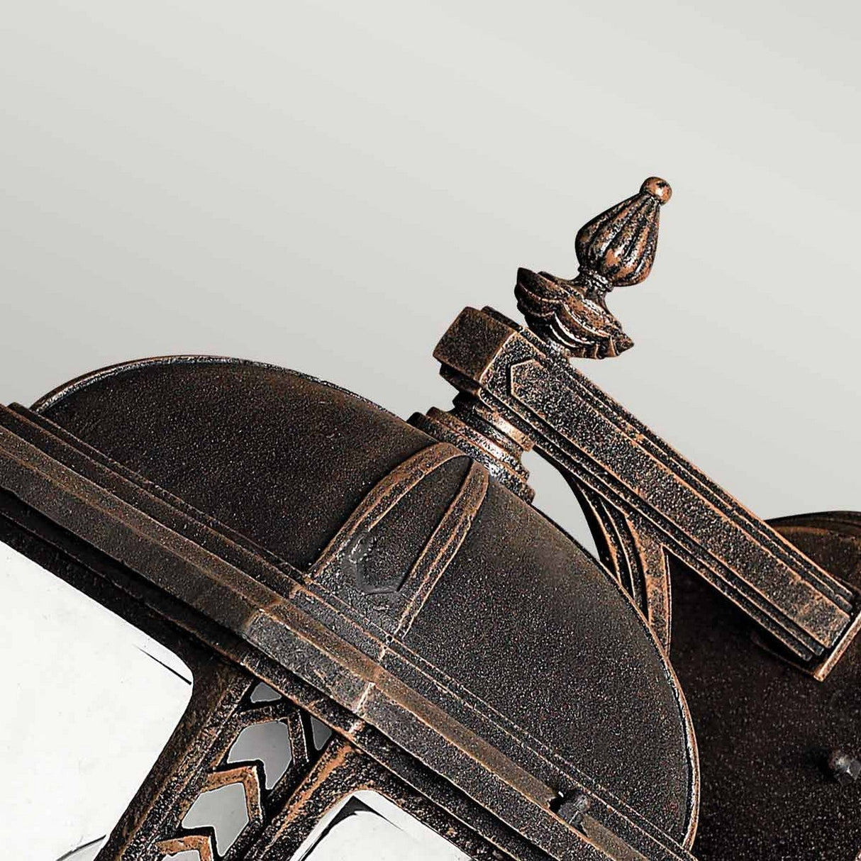 Close-up of a decorative Wabash Outdoor 2 Light Medium Wall Lantern in a bronze finish. The lantern showcases intricate details, such as a decorative finial on top and chevron patterns along the edges. The background is plain and light-colored.