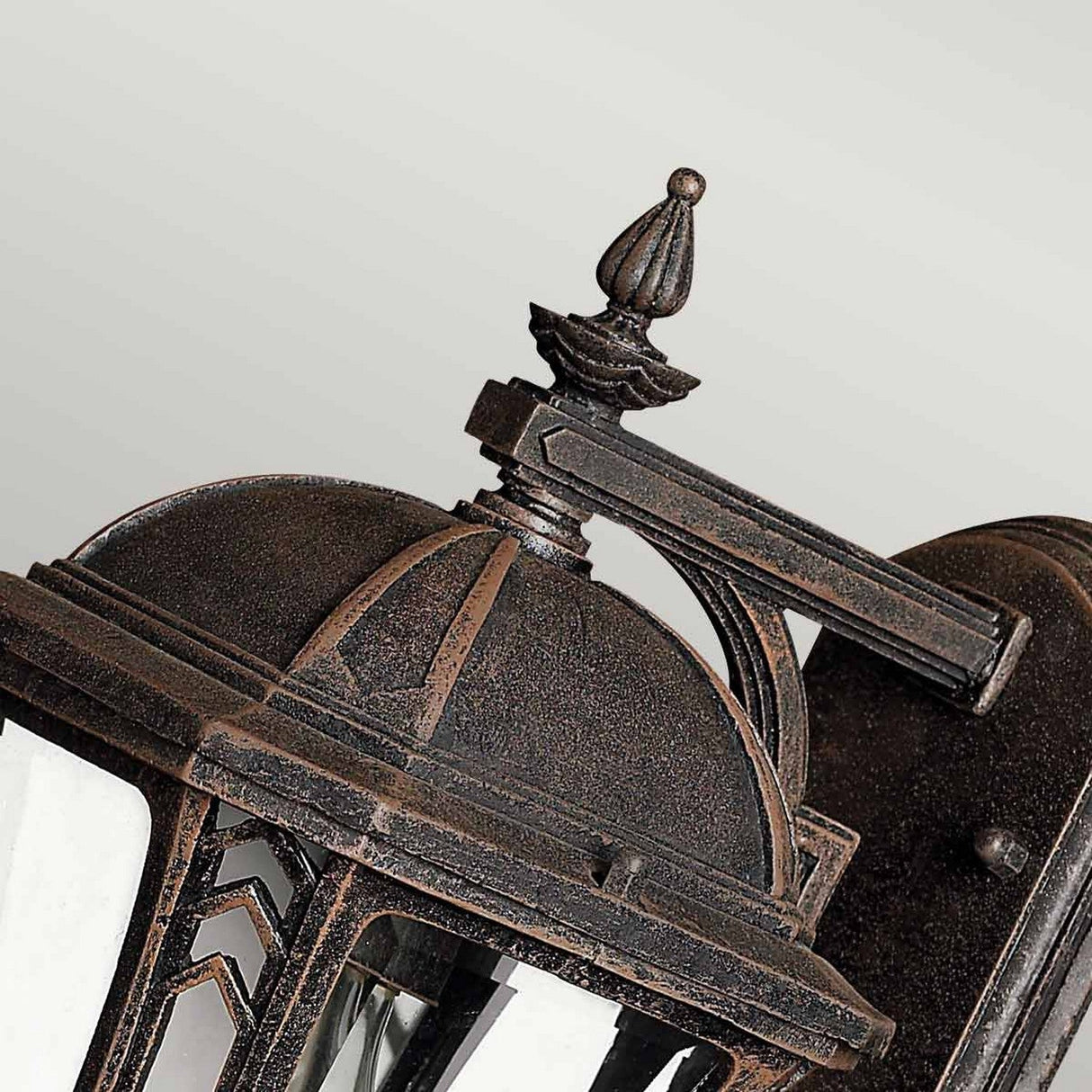 Close-up of the Wabash Outdoor 1 Light Small Wall Lantern in bronze, showcasing intricate chevron pattern detailing. It features a dome-shaped top and decorative finial with clear glass panels and an ornate mounting arm, set against a light gray background, making it perfect for enhancing your outdoor lighting.