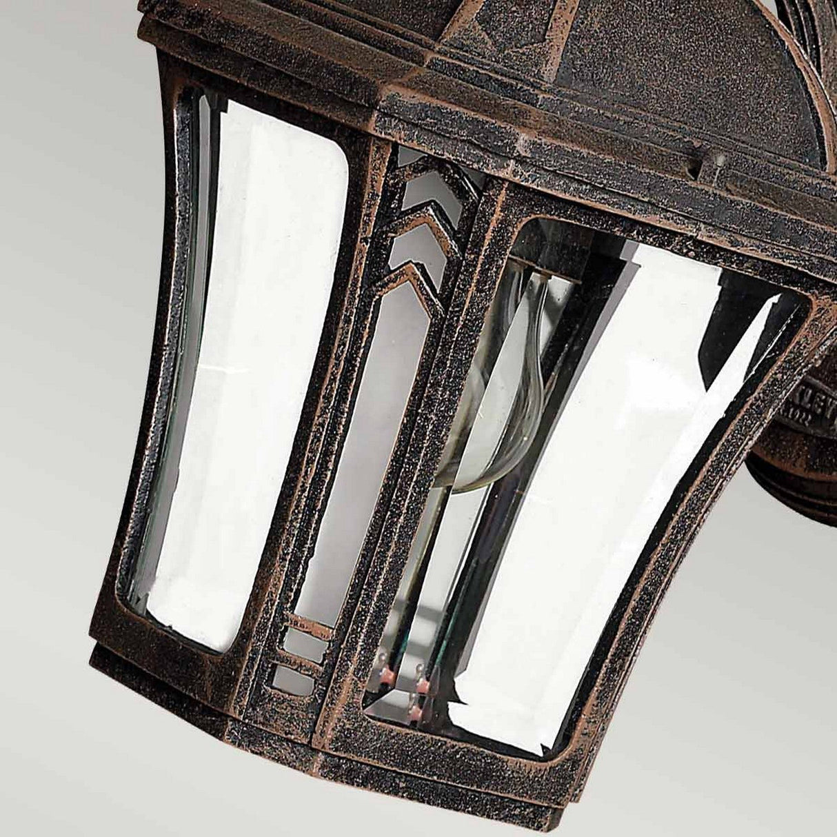 A close-up of the Wabash Outdoor 1 Light Small Wall Lantern in bronze highlights its clear glass panes, revealing the visible light bulb within. This outdoor lighting piece features a rustic, weathered finish with ornate detailing and subtle hints of a chevron pattern.