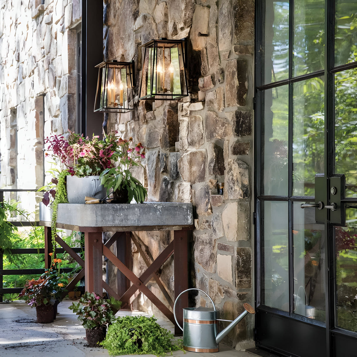 A picturesque outdoor setting showcases a stone wall highlighted by the Walker Outdoor 2 Light Large Wall Lantern in Bronze with Heritage Brass. Two of these charming glass lanterns cast a warm glow, while a metal planter brimming with vibrant greenery is perched on a wooden stand next to a watering can. The glass door mirrors the surrounding plants, enhancing the serene atmosphere with sophisticated outdoor lighting.