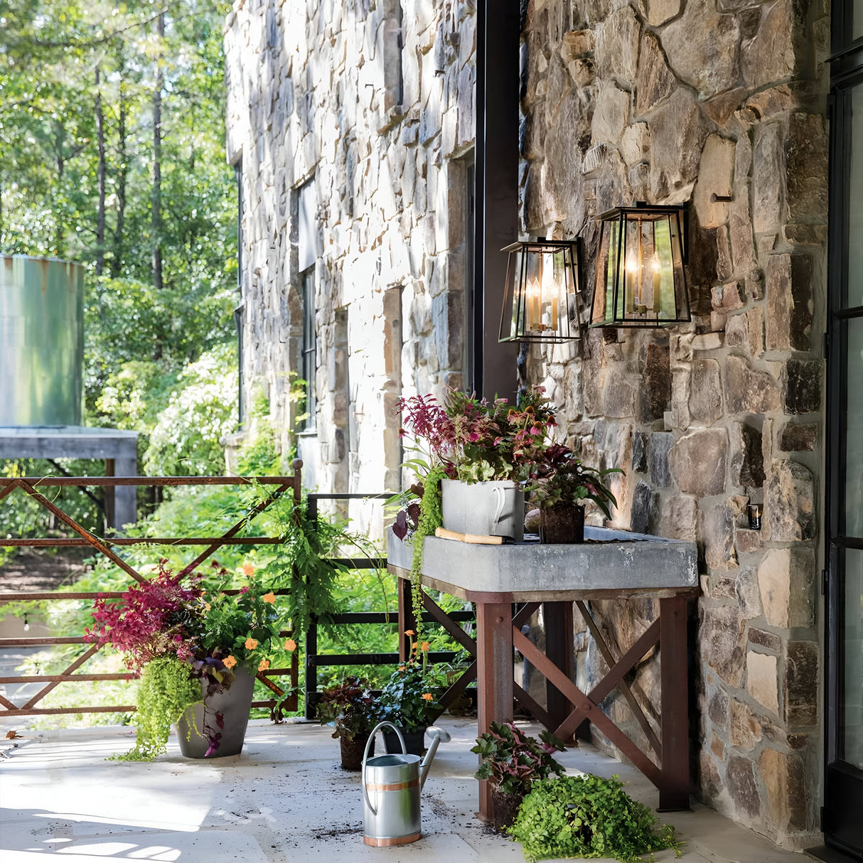 A rustic outdoor patio boasts a stone wall decorated with the Walker Outdoor 2 Light Large Wall Lantern in Bronze with Heritage Brass. This dual-light arrangement casts a warm glow over a table displaying a tub filled with an assortment of plants, encircled by additional green foliage. A watering can lies on the ground, while trees offer a lush backdrop.