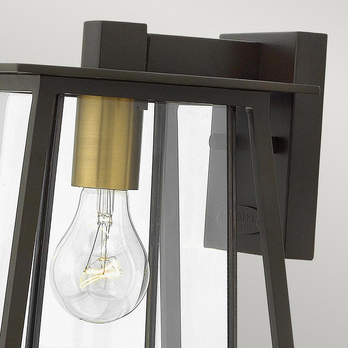 Close-up of the Walker Outdoor 1 Light Small Wall Lantern in bronze with a heritage brass finish, showcasing a clear glass panel and a visible light bulb inside. The fixture has a rectangular shape with a brass interior, mounted on a flat square base against a light gray background. Ideal for adding elegance to outdoor lighting.