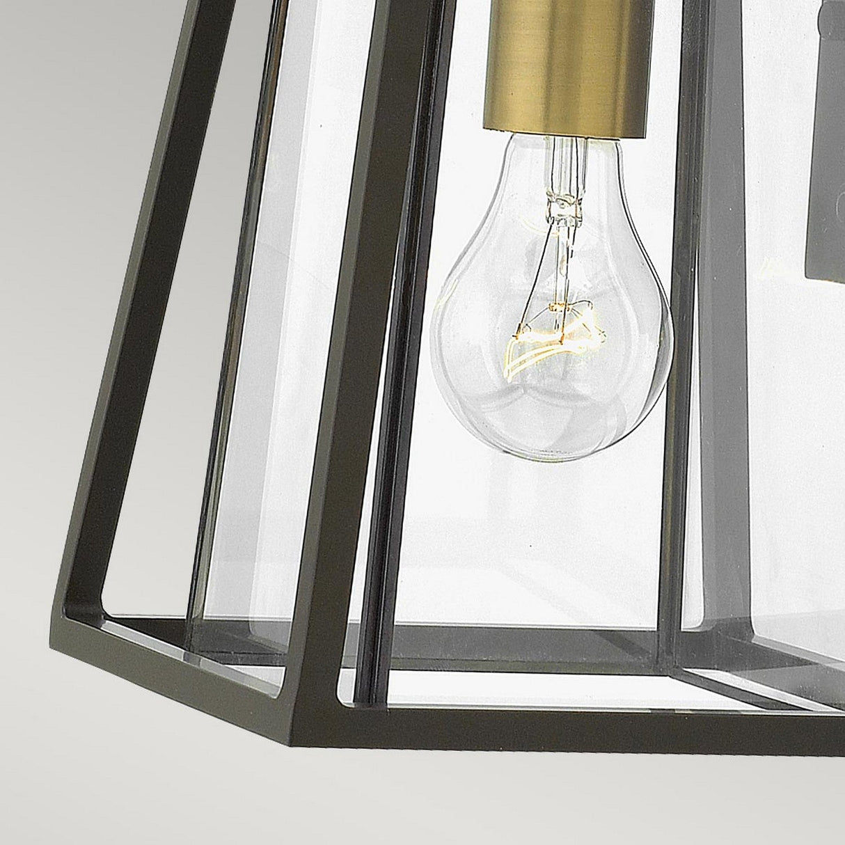 The Walker Outdoor 1 Light Small Wall Lantern in Bronze with Heritage Brass features a geometric metal frame with a clear glass enclosure, elegantly showcasing an exposed bulb. Its sleek, modern lines set against a simple backdrop exude sophistication, akin to a contemporary bronze wall lantern.