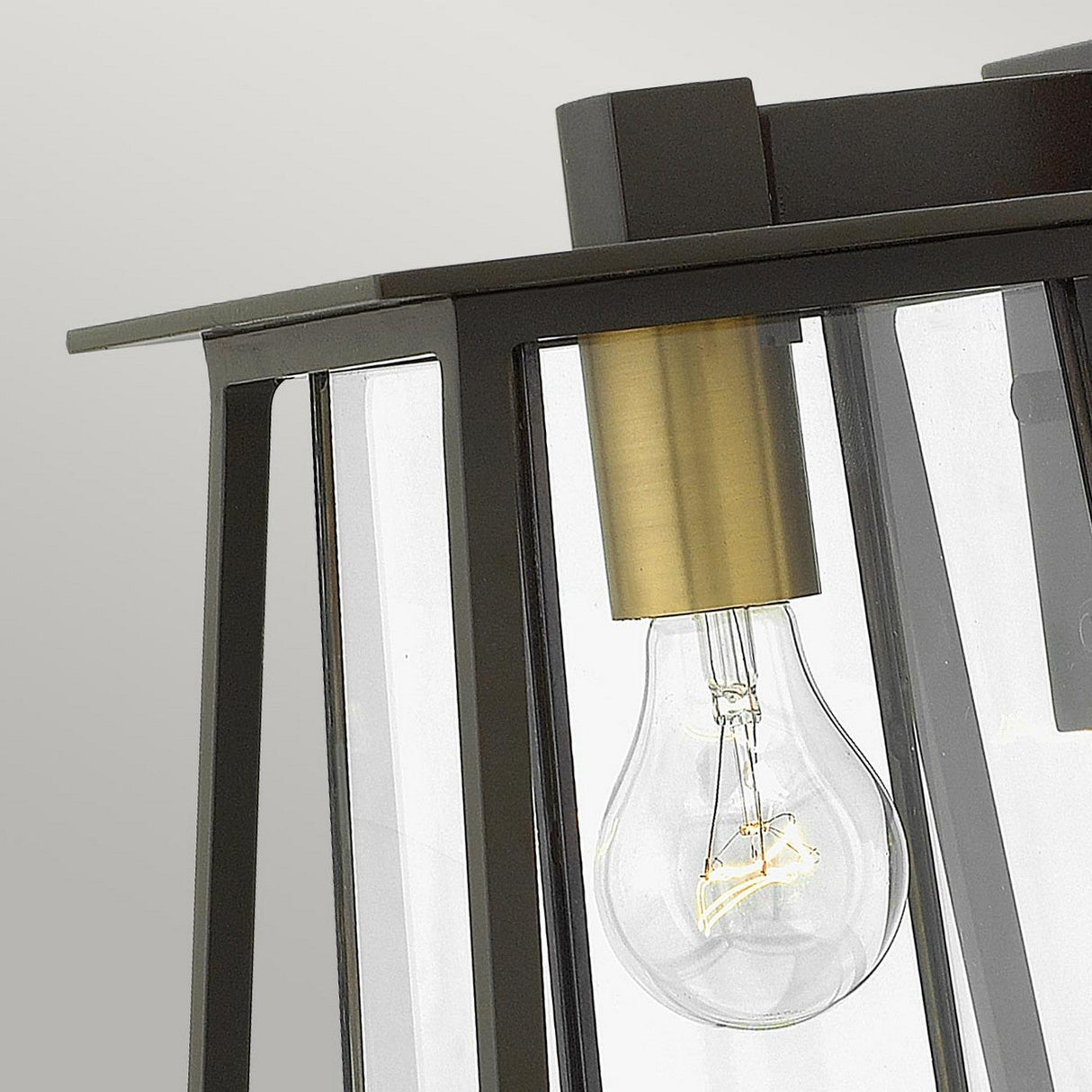 A detailed view of the light bulb within the Walker Outdoor 1 Light Small Wall Lantern in bronze with heritage brass showcases its elegant design. The matte-finished metal encases the bulb, which is visible through clear glass panels, set against a plain, light background that complements sophisticated outdoor lighting.