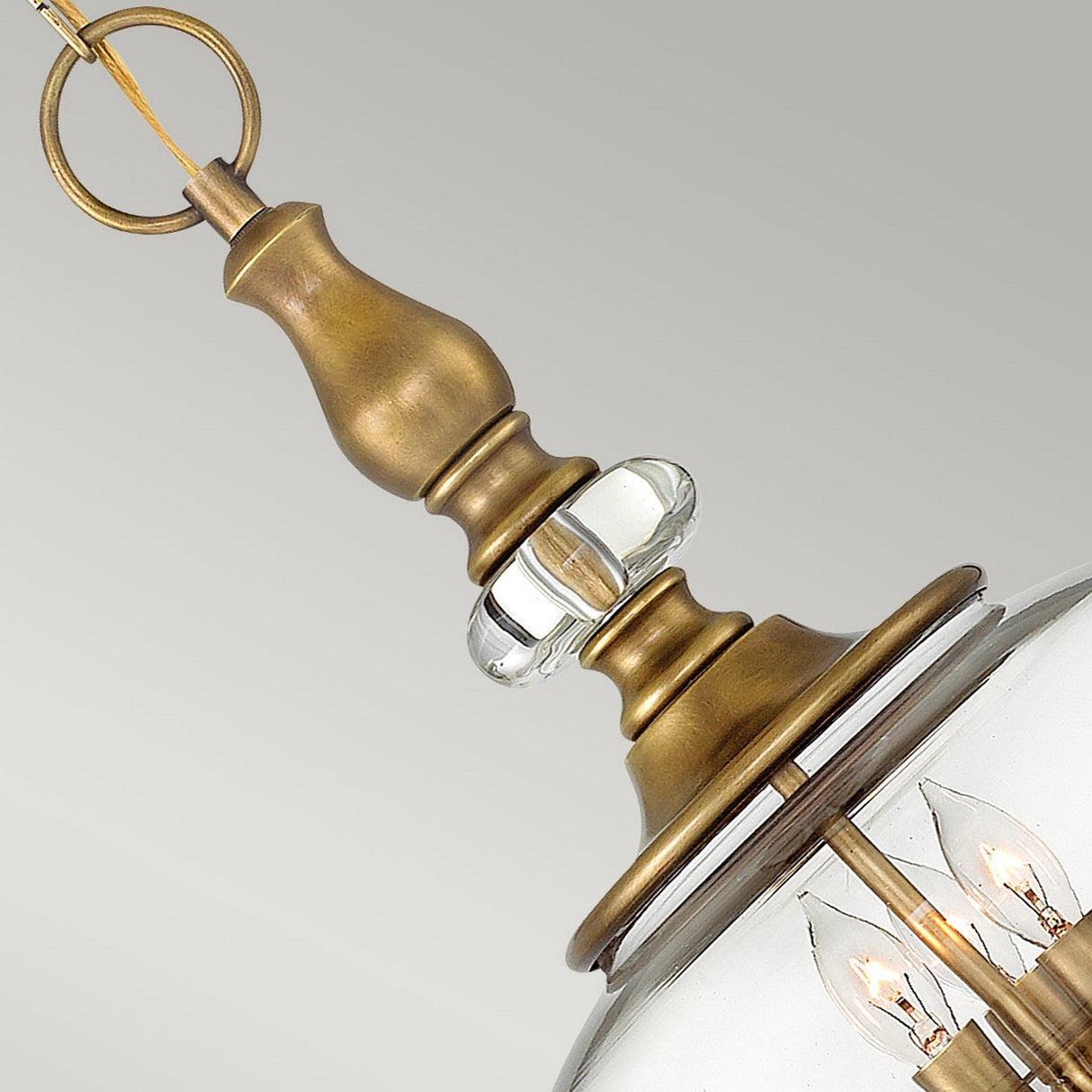 Close-up of the Wexley 3 Light Chandelier in Heritage Brass, showcasing a brushed brass stem with a clear glass accent and supporting a dome-shaped glass shade. Inside, candle-style bulbs provide soft illumination against a gentle gray background for a seamless transitional design.