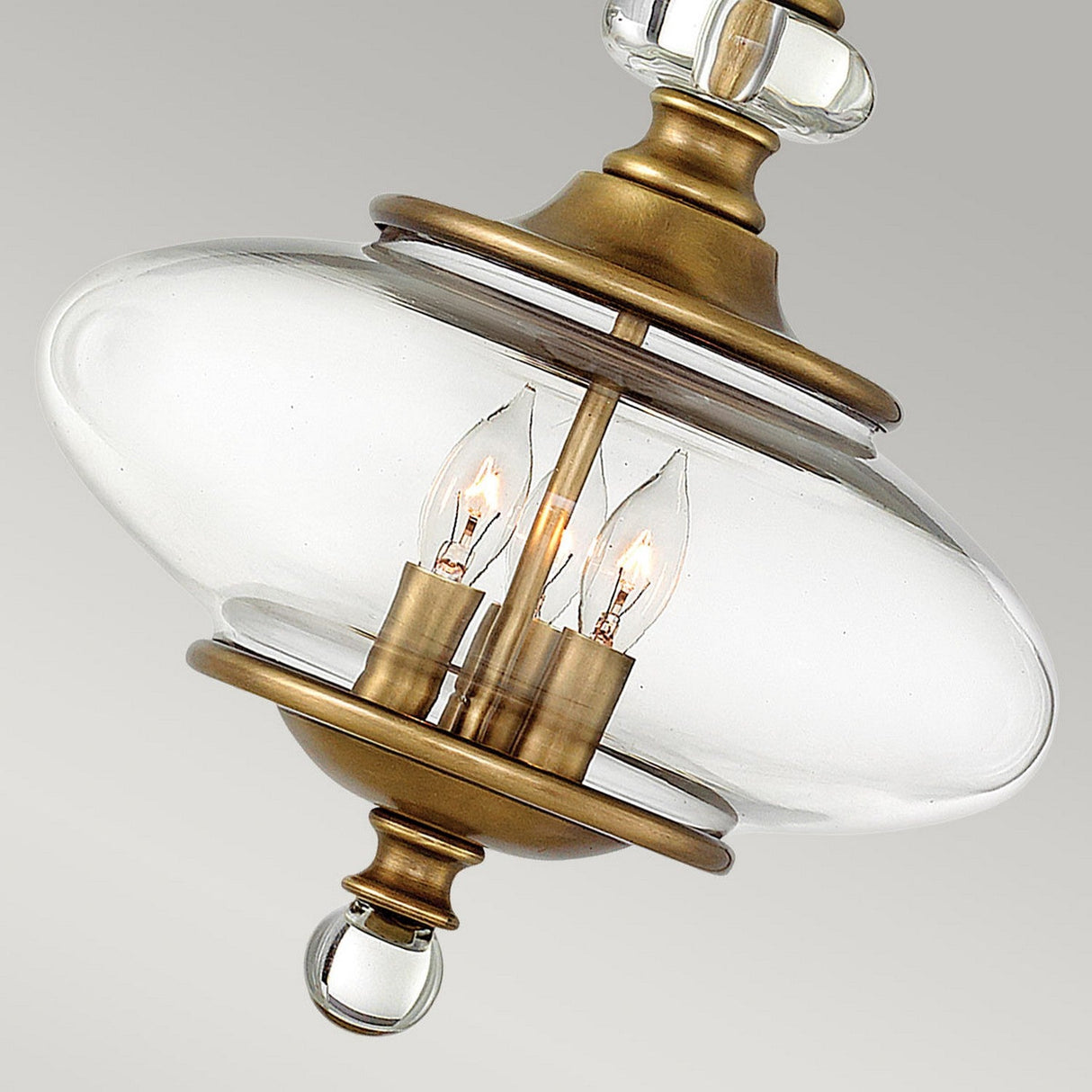 A close-up of the Wexley 3 Light Chandelier - Heritage Brass showcases an elegant ceiling fixture with a Heritage Brass finish and clear glass design. It features three exposed candle-like bulbs inside a round glass enclosure, hanging against a light gray background, highlighting its transitional design.