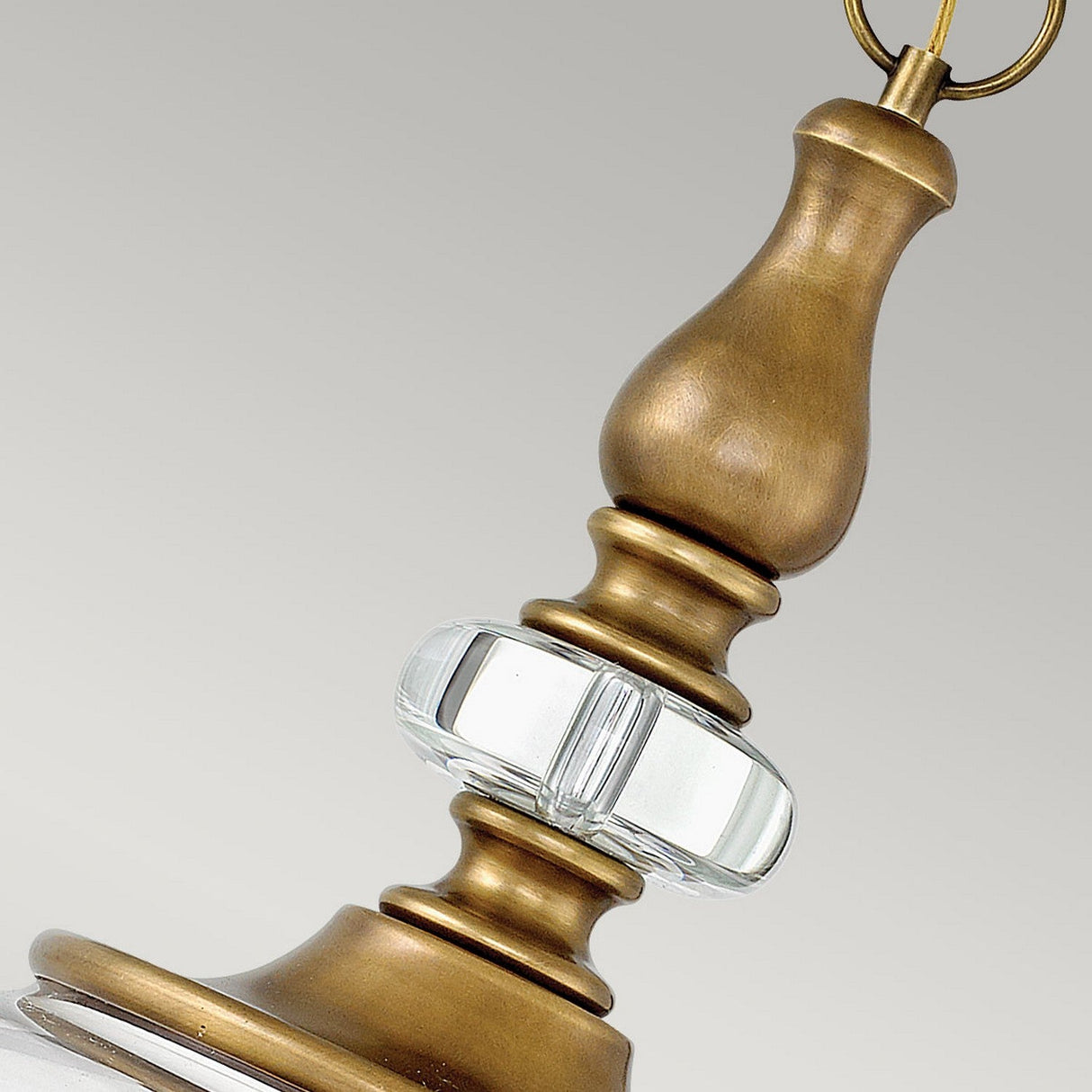 A close-up of the Wexley 5 Light Chandelier in Heritage Brass showcases its vintage-inspired elegance. The fixture features a clear glass centerpiece and an ornate, tapered design, hanging gracefully from a circular hook against a neutral background.