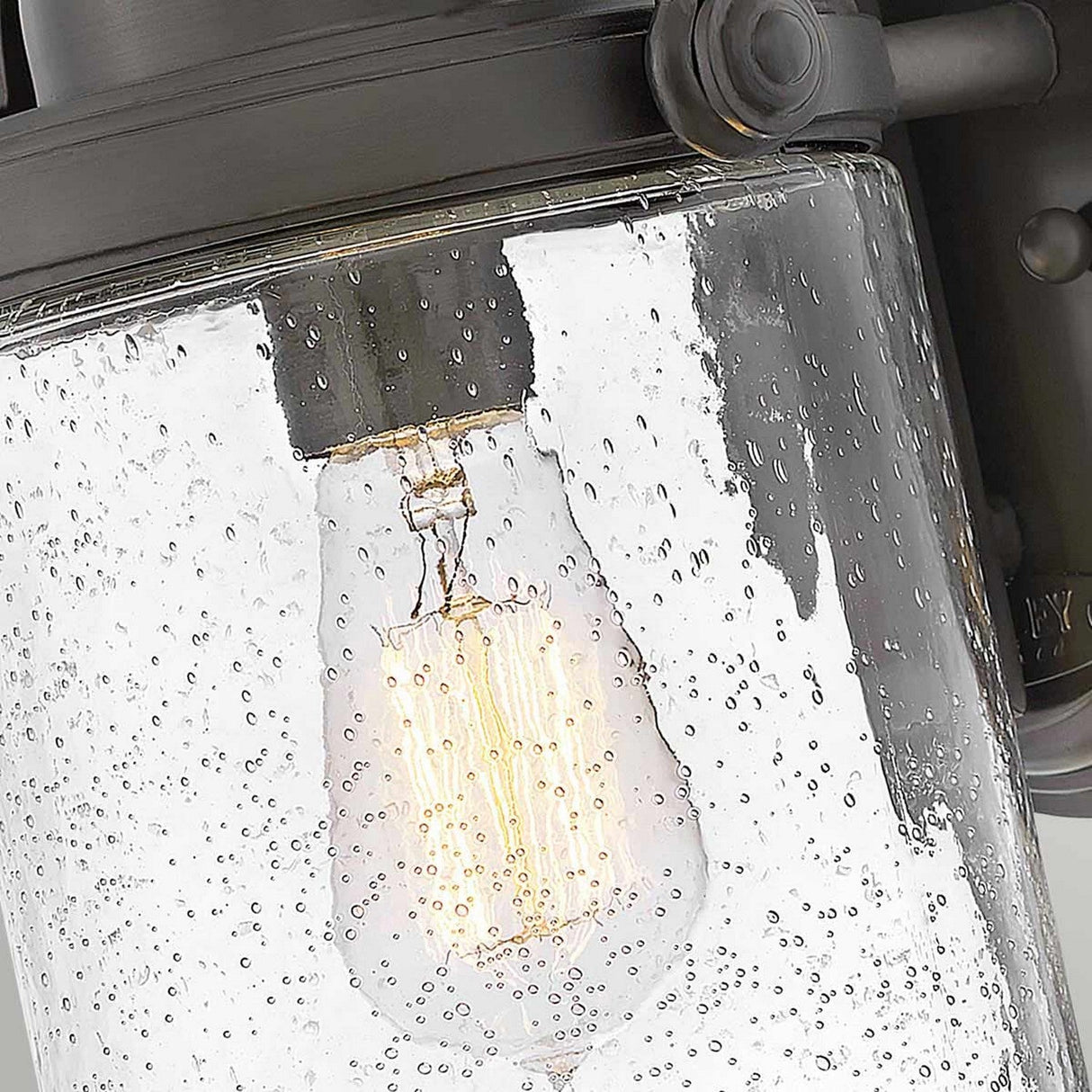 A close-up of the Willoughby 1 Light Wall Lantern reveals a vintage-style light bulb glowing inside a cylindrical glass fixture. The bubbled surface amplifies the warm, ambient glow. Constructed with oil rubbed bronze, this lantern infuses outdoor lighting with an industrial flair and dark metal charm.
