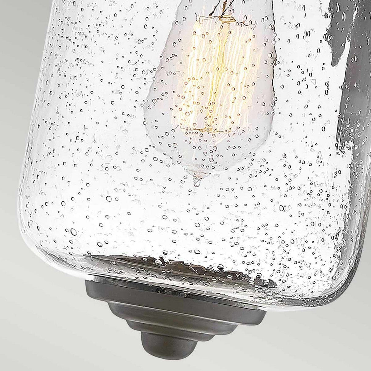 A close-up of the Willoughby 1 Light Wall Lantern in oil rubbed bronze reveals a light bulb inside a clear glass fixture with small embedded bubbles that create a textured appearance. The softly glowing bulb casts light through the patterned glass, reminiscent of an elegant outdoor lantern.