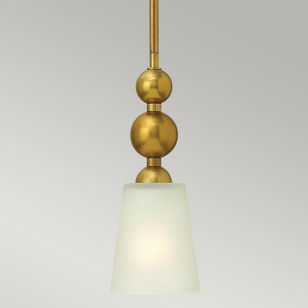 The Zelda 1 Light Mini Pendant in a vintage brass finish features a gold metal rod and two spherical accents. Its mid-century design includes a frosted white, cone-shaped glass shade that casts a soft, diffuse glow against the plain gray background.