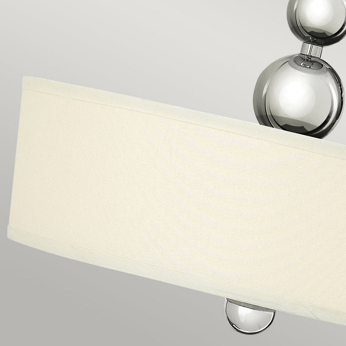 A close-up of a contemporary lamp radiates mid-century appeal with its beige fabric drum shade. The polished nickel base highlights silver metallic spheres, positioned at the top and below the shade, reminiscent of the Zelda 3 Light Pendant - Polished Nickel against a neutral gray backdrop.