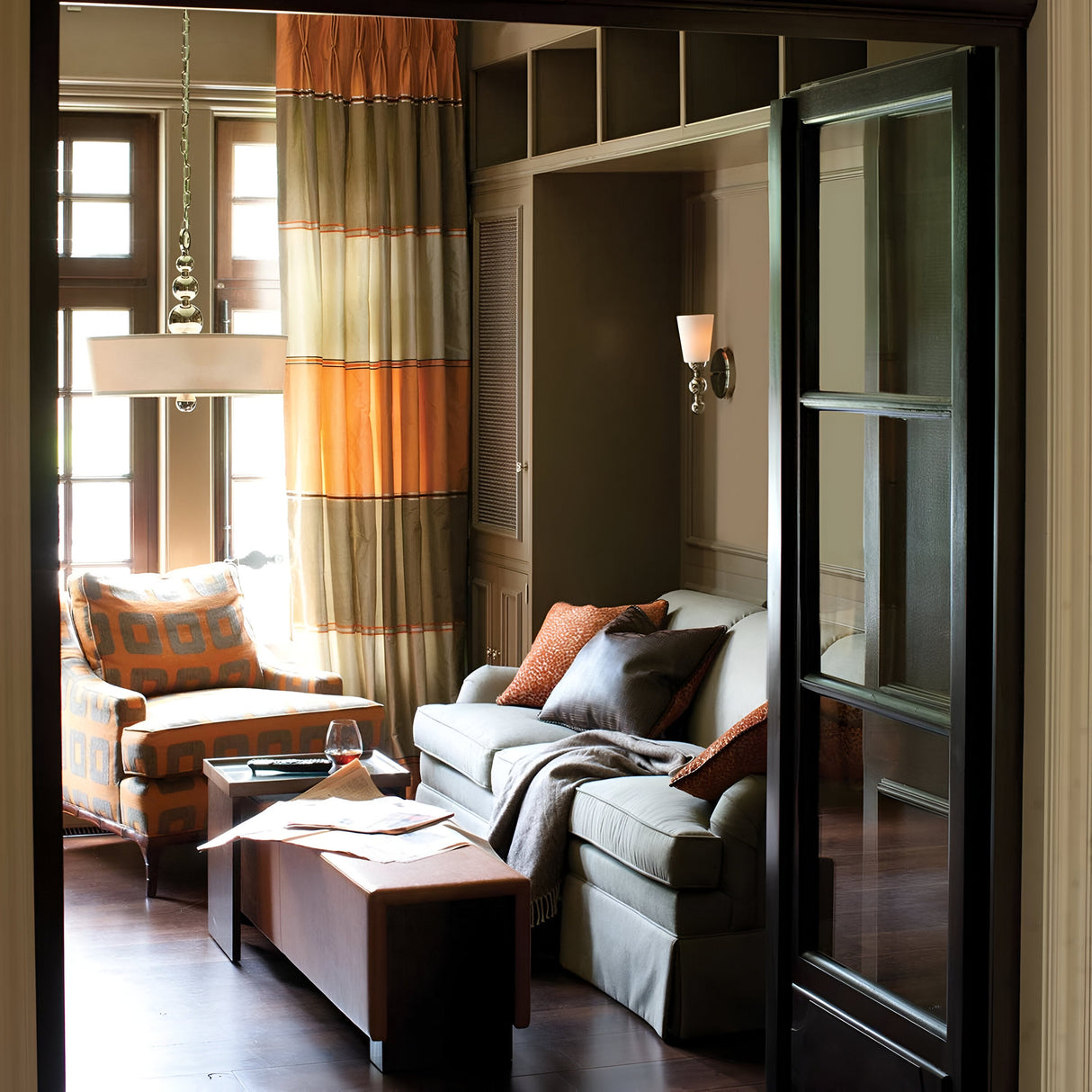 The inviting living room features a sofa, an armchair, and a cushioned coffee table. The mid-century charm is accentuated by earthy tones in the orange and brown cushions and curtains. Natural light pours in through large windows, complemented by the elegant touch of the Zelda 3 Light Pendant in Polished Nickel, enhancing the warm ambiance.