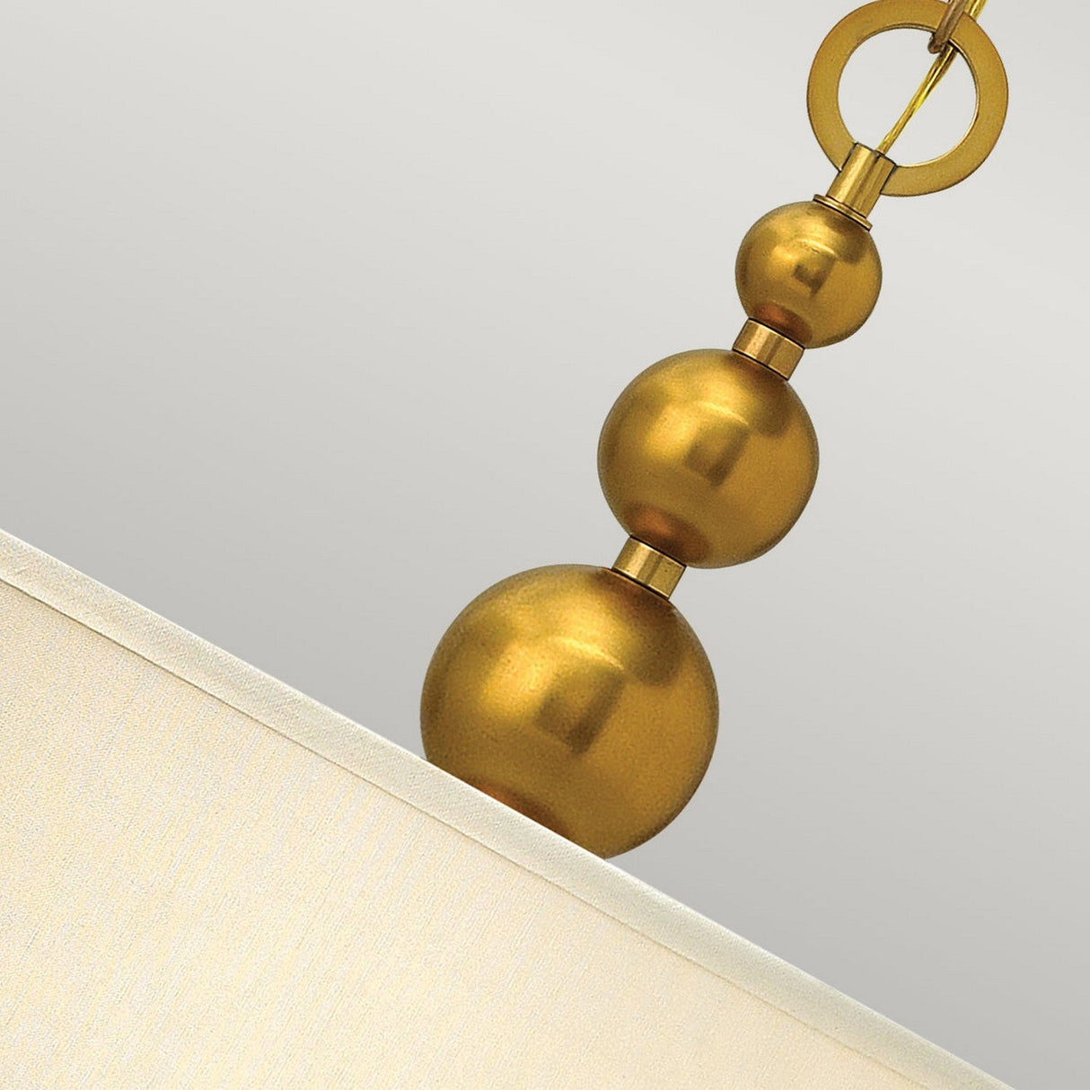 A close-up of the Zelda 3 Light Pendant - Vintage Brass showcases a modern fixture that elegantly merges mid-century design with vintage brass accents. It displays three metallic gold spheres in descending sizes, seamlessly linked vertically with a metal ring at the top, while a fabric lampshade is partially visible in the corner.