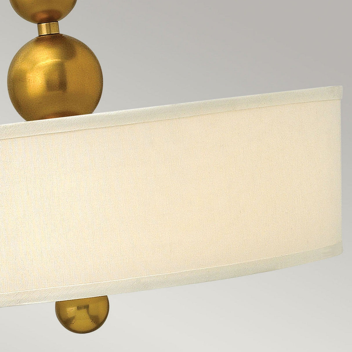 The Zelda 3 Light Pendant - Vintage Brass showcases a sleek, mid-century aesthetic with its two stacked vintage brass spheres positioned above and below a prominent cylindrical beige fabric lampshade. The elegant design is further highlighted against a soft gray background.