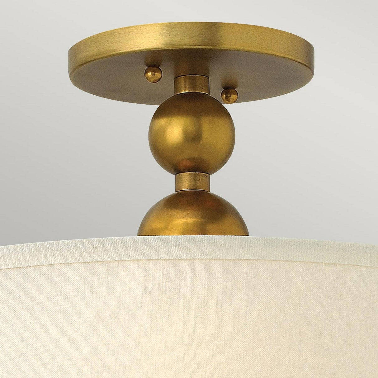 Close-up of the Zelda 3 Light Semi Flush - Vintage Brass, showcasing a mid-century design with a gold, round base and decorative features. Below it hangs a white, cylindrical lampshade against a plain light gray background.