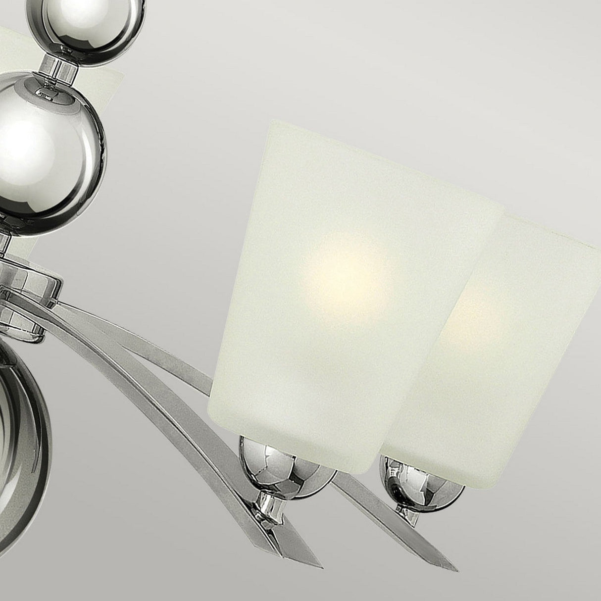 A close-up of the Zelda 5 Light Chandelier - Polished Nickel showcases two white frosted glass shades, each illuminated by a glowing bulb. The fixture embodies mid-century sophistication with its sleek, curved metallic arms and polished nickel accents, set against a soft gray background.
