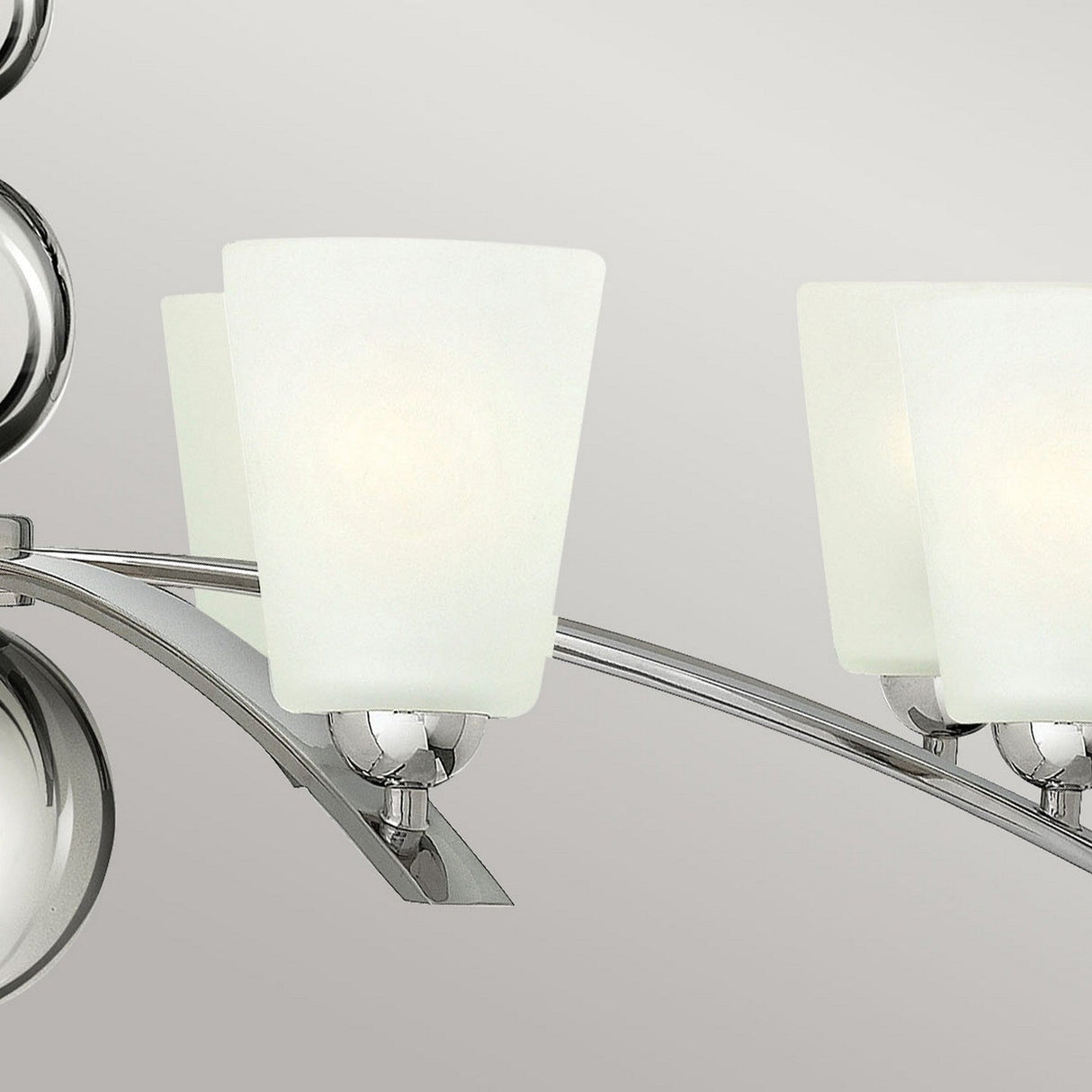 Close-up of the Zelda 8 Light Chandelier in a polished nickel finish, showcasing its modern design inspired by mid-century aesthetics. The chandelier features frosted white glass shades in square shapes with rounded edges, all elegantly attached to a curved metal arm. The backdrop is a neutral gray.